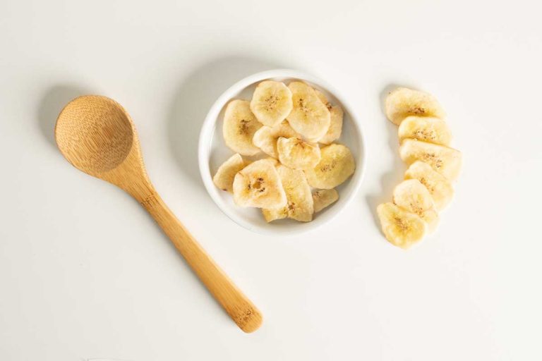 Dried banana slices in a small white bowl with a wooden spoon to the left hand side.