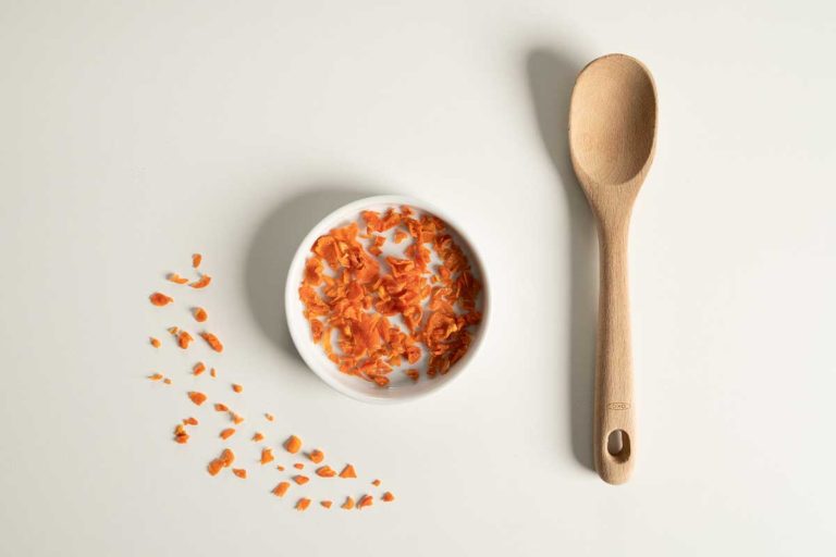 Dehydrated cut carrots in a white bowl with a wooden spoon to the right hand side.
