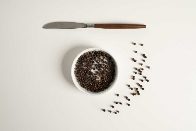 Whole black pepper in white bowl with flat butter knive.