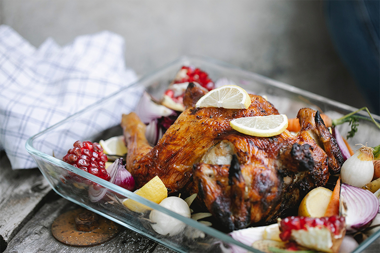 Whole chicken on a glass baking tray surrounded by onion, lemon, and pomegranate.