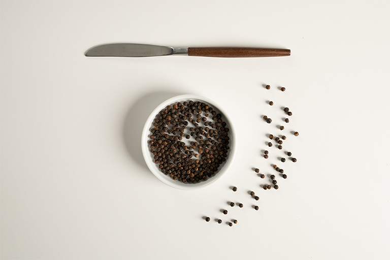 Whole black pepper balls in a small white bowl with a knife over it and whole round pepper balls next to it.