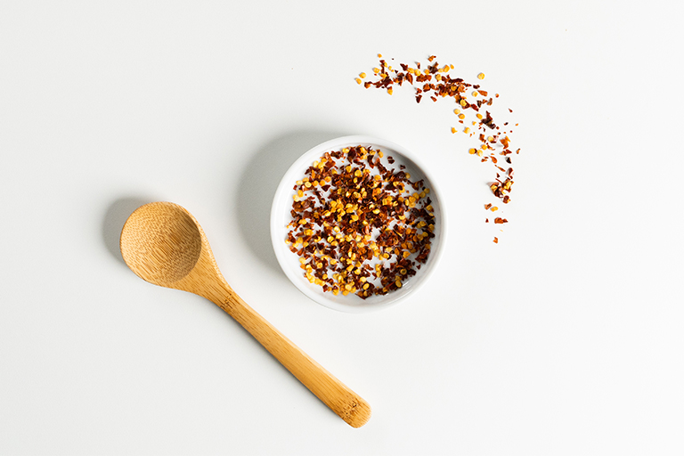 On the left side of a small white bowl with ground crushed red pepper their is a wooden spoon.