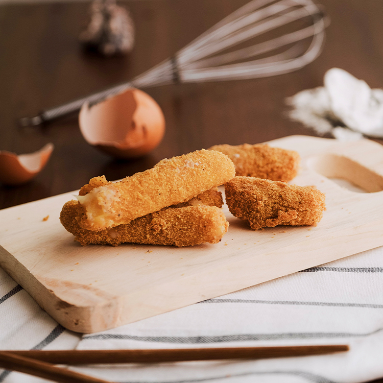 breaded mozzarella sticks on wooden cutting board with broken egg and whisk blurred in the background
