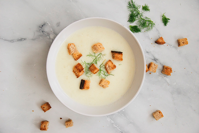 Yellowish Soup with croutons and fresh dill on top in a white bowl on top of a marble countertop