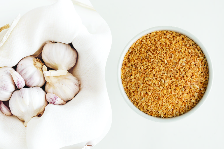 group of garlic head in a towel in basket next to photo of dehydrated garlic in bowl white bowl on a white countertop