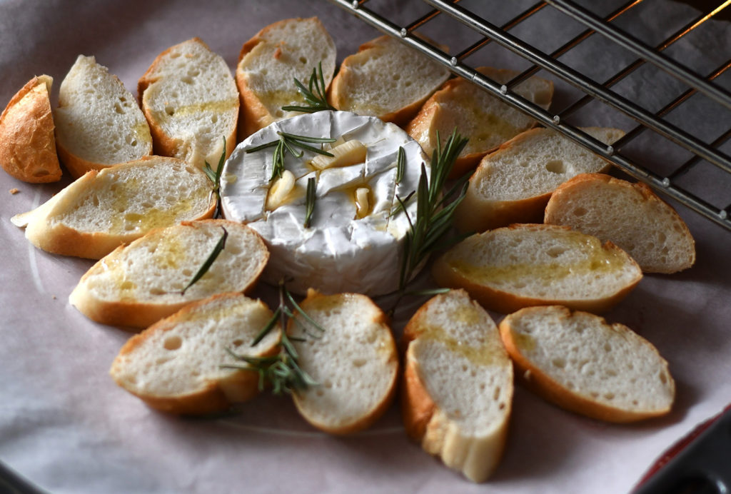round white Cheese with whole garlic cloves stuffed into the middle and slices of bread surrounding it with a drizzle of oil and sprinkle of fresh rosemary