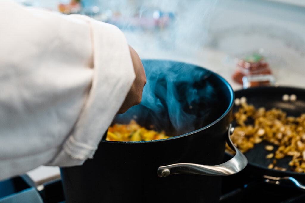 A chef preparing the smoky Ratatouille soup in a large black pot on a stove and steaming rising - page out of the measures cookbook