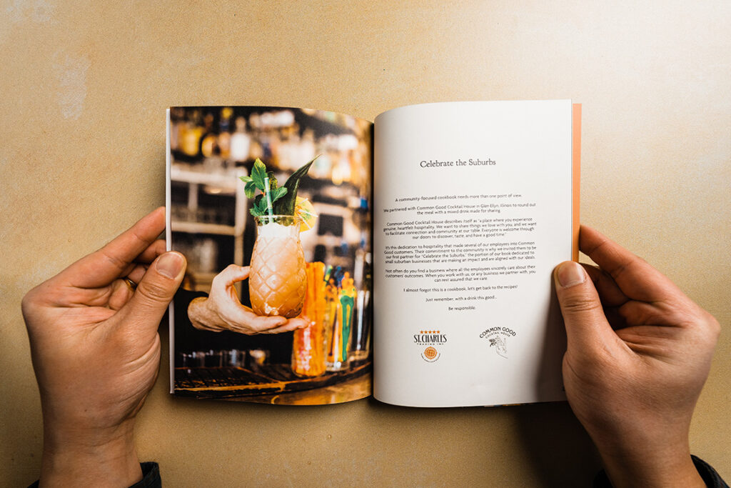 hands holding a book open to a page with an image of a hand holding a pineapple glass drink and a page titled Celebrate the Suburbs