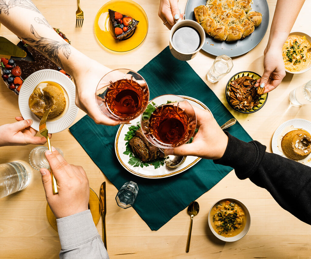 two hands toasting glasses in the middle of a table while other hands are grabbing for food.