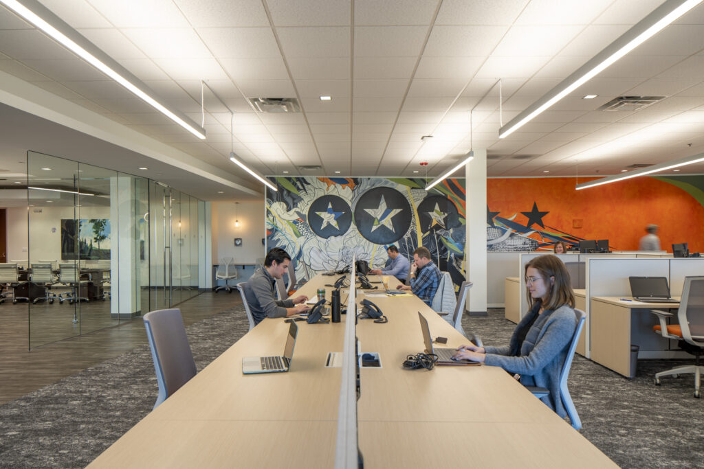 Long table office setting with four people working at laptops as food ingredient distributors; open cubicles on the right and glass wall on the left.