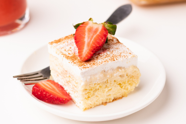 A delicious tres leches on a white plate, topped with a red strawberry on top.