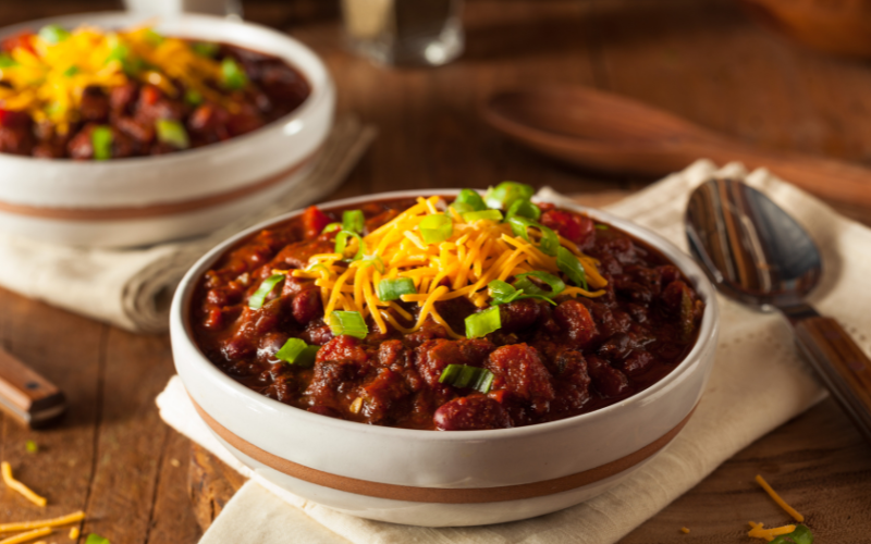 Two chili dishes on top of a wooden table with napkins rolled underneath and spoons to the side.