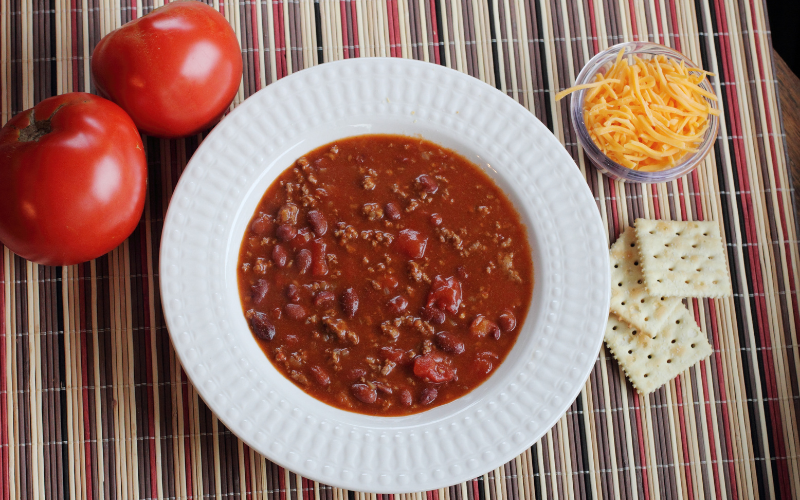 Classic chili dish on a striped mantel, surrounded by tomatoes, cheese, and crackers.
