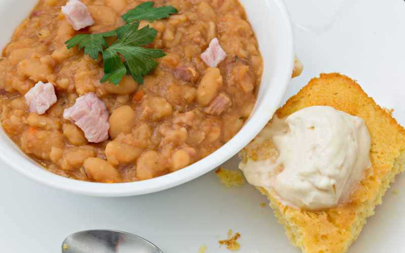 Mushed navy beans mixed with chicken in a white bowl on top of a white surface. To the right sits a slice of cornbread.