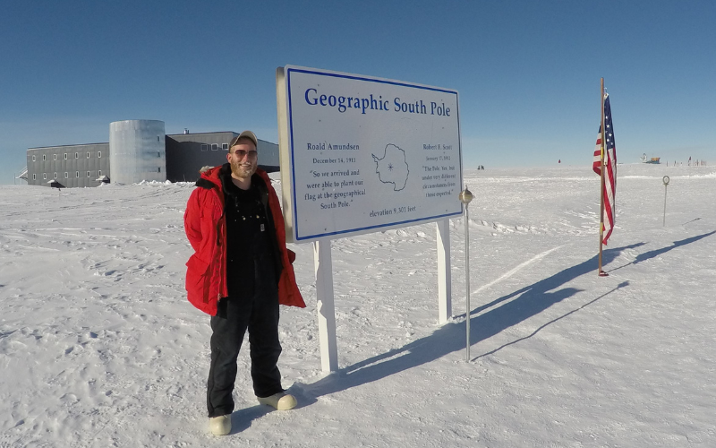 Brian Eisenstatt standing next to the geographic South Pole marker in Antarctica