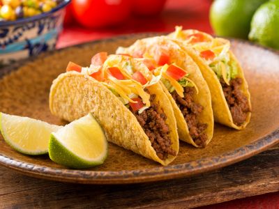 A photo of three hard shelled tacos with chili powder and limes on the side on a copper plate.