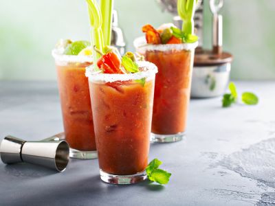 A photo of three Bloody Mary cocktails with celery salt rims and vegetable garnishes sitting on a grey countertop.