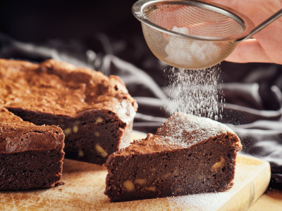 A photo of a brownie slice being covered in powdered sugar. It is sitting on a wooden cutting board.