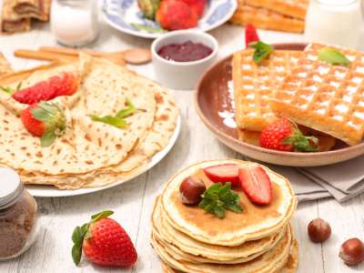 A photo of pancakes, waffles, and crapes, all on separate plates with berries on a white wood table.