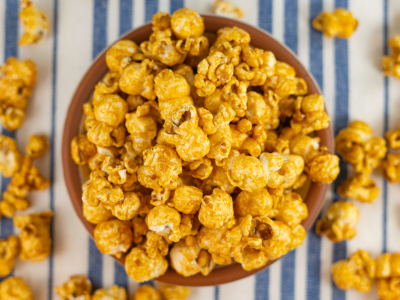 A photo of popcorn in a bowl with popcorn surrounding the bowl. The popcorn is yellow with seasoning.