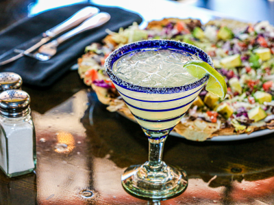 A photo of a margarita in a blue and clear glass with the rim salted and a lime. In the background there's a plate of nachos.