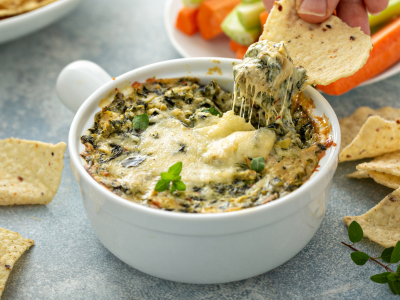 A photo of spinach dip in a white ceramic bowl. A person is dipping a chip into the dip.
