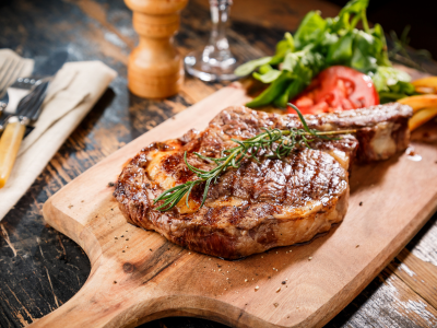 A photo of a grilled steak on a wooden cutting board with tomatoes and lettuce on the side sitting on a wooden table.