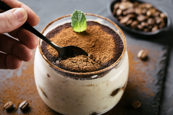 A photo of a clear glass cup filled with milk tea tiramisu and someone is taking a spoon full of it. The glass is on a black countertop with coffee beans in the background.