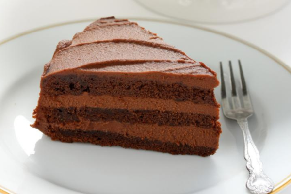 A photo of a slice of chocolate cake sitting on a white plate with a silver fork.