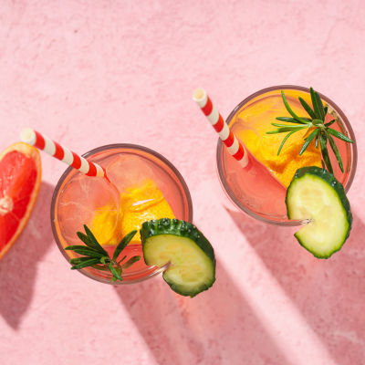 A photo of two pink drinks with grapefruit and cucumber garnishes. They are sitting on a pink surface.