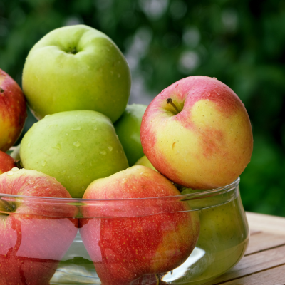 A photo of a bowl of green and red apples in a clear bowl sitting outside on a wooden table.