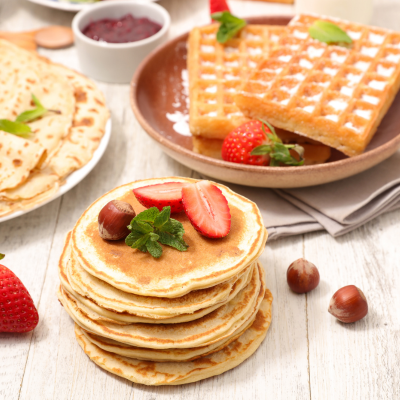A photo of a stack of pancakes topped with strawberries. In the background there's a plate of waffles and a plate of crepes.