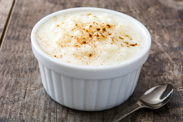 Classic creamy rice pudding with cinnamon in a white container, silver spoon diagonally bottom right, on a brown-grayish wooden surface