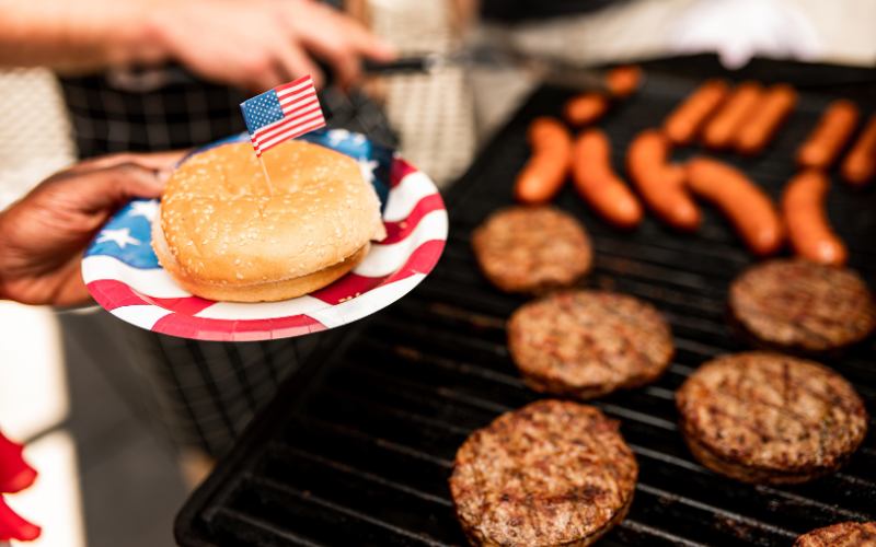 A burger on a 4th of july themed plate next to a grill firing up some patties for Independence Day