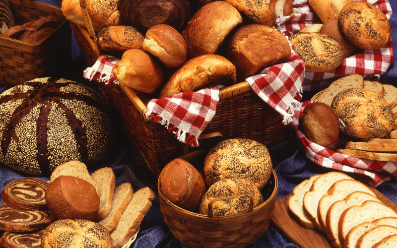 Bread basket full of different rolls surrounded by blue and red tablecloths on a table full of more types of bread